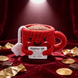 May include: A red crocheted tea cup with a smiling face and a tea bag. The cup holds a brown substance, possibly tea. A white sign reads "POSITIVI-TEA" with an inspirational message. Gold coins and chocolate pieces are scattered around.