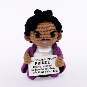 Peut inclure: Poupée au crochet représentant Prince, avec une afro noire, une peau brune et une veste violette. La poupée tient une pancarte avec le texte "MOTIONAL SUPPORT PRINCE Dearly beloved, I'm here to get thro this thing called life."