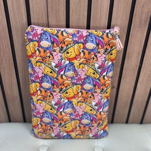 Op de afbeelding: Een rechthoekig etui met een kleurrijk patroon van stripfiguren, waaronder Winnie de Poeh, Iejoor, Knorretje en Teigetje. Het etui heeft een roze rits met een kwastje en staat tegen een houten achtergrond.