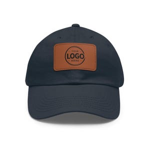 May include: A navy blue baseball cap featuring a rectangular brown leather patch centered on the front. The patch displays a circular graphic with the text YOUR LOGO HERE printed in black. The cap has a curved brim and stitched eyelets.