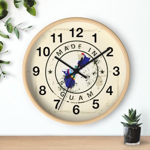 May include: A round wall clock with a light wood frame. The clock face is cream-colored with black numerals and hands. The center features a map of Guam and the text "MADE IN GUAM".