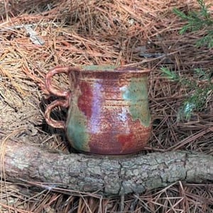 Könnte beinhalten: Eine handgefertigte Keramiktasse mit zwei Henkeln. Die Tasse hat einen braunen Boden mit roten und grünen Akzenten. Die Tasse steht auf einem Holzstück, umgeben von Kiefernnadeln und Blättern.