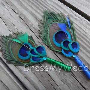 May include: Two peacock feather boutonnieres with blue and green accents. The feathers are arranged in a circular pattern and are attached to a blue and green ribbon.