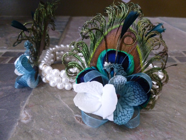 May include: A pair of decorative accessories featuring white pearl bracelets, peacock feathers, and fabric flowers. The flowers are white and shades of blue, with blue ribbon accents. The accessories are arranged on a stone surface.
