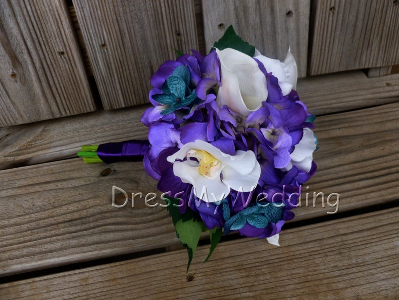 May include: A purple and white bridal bouquet with hydrangeas, orchids, and calla lilies. The bouquet is tied with a purple ribbon and has green leaves.