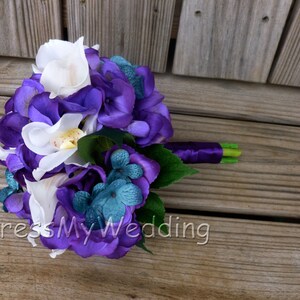 May include: A purple and white bridal bouquet with white orchids, purple hydrangeas, and teal blue flowers. The bouquet is tied with a purple ribbon.