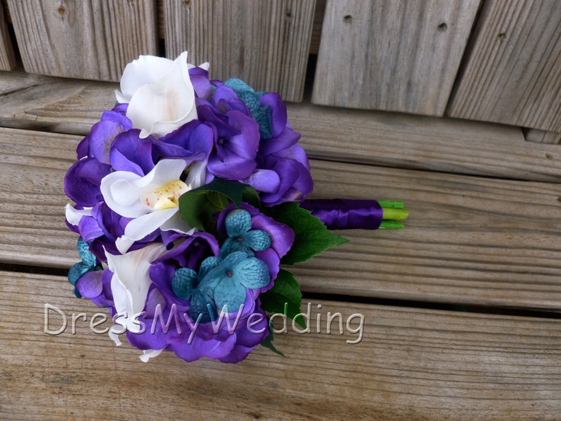 May include: A purple and white bridal bouquet with white orchids, purple hydrangeas, and teal blue flowers. The bouquet is tied with a purple ribbon.