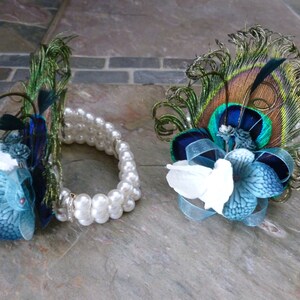 May include: Two decorative floral accessories. One is a bracelet with white pearls and teal and white flowers. The other is a floral pin with peacock feathers, teal and white flowers, and blue ribbon.
