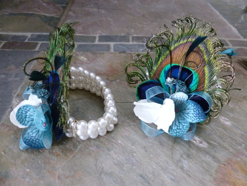 May include: Two decorative floral accessories. One is a bracelet with white pearls and teal and white flowers. The other is a floral pin with peacock feathers, teal and white flowers, and blue ribbon.