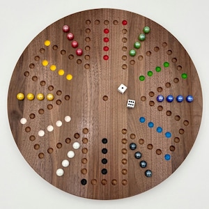 Può includere: Un gioco da tavolo rotondo in legno con finitura marrone scuro. Il tabellone presenta fori e biglie colorate in giallo, rosso, verde, blu, bianco e nero. Un dado bianco è al centro.