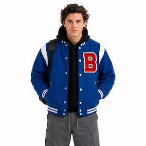 Può includere: Una giacca varsity blu con maniche bianche, polsini e orlo a coste con righe bianche e una grande toppa con la lettera B rossa e bordo giallo sul petto. La giacca presenta bottoni a pressione argentati sul davanti ed è indossata sopra una felpa nera.