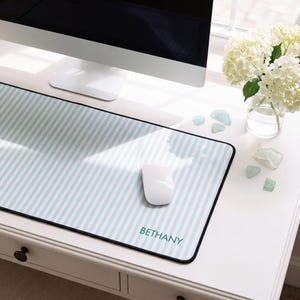 May include: A rectangular desk mat featuring a light blue and white vertical striped pattern with the name BETHANY printed in dark teal lettering. The mat has a black border and a smooth surface, with a white computer mouse resting on top.