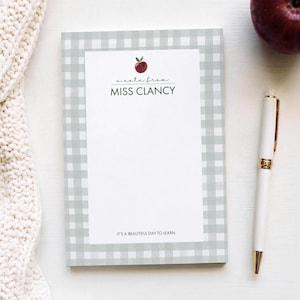May include: A rectangular notepad with a light gray and white gingham border. The center features a red apple illustration, the text "a note from MISS CLANCY" in black, and the phrase "IT'S A BEAUTIFUL DAY TO LEARN" at the bottom.