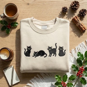 May include: Cream-colored sweatshirt featuring embroidered black cat designs. The cats are in various poses. The sweatshirt is surrounded by holiday-themed decorations, including holly, pine cones, and cinnamon sticks.