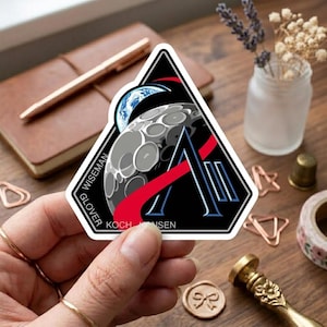 May include: A triangular sticker featuring a stylized design of the moon, Earth, and a large letter "A" with red and blue accents. The sticker includes the names "Wiseman", "Glover", "Koch", and "Hansen".