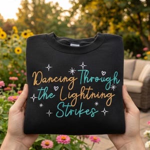 May include: Black sweatshirt with the embroidered phrase "Dancing Through the Lightning Strikes" in gold, teal, and white. The design includes stars and hearts. The sweatshirt is held up in front of a garden.