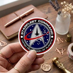 May include: A round sticker featuring the Starfleet Command emblem from Star Trek. The design includes a silver arrowhead shape with a black star, a red swoosh, and the words "STARFLEET COMMAND" and "UNITED FEDERATION OF PLANETS" in white text on a navy blue background.