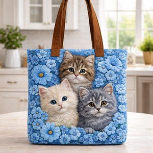 Może przedstawiać: Niebieska torba z kwiatami i trzema kociętami. Torba ma brązowe uchwyty i kwadratowy kształt. Kocięta mają brązowe, białe i szare futro. Idealna dla miłośników kotów.