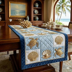 Pode incluir: Caminho de mesa acolchoado azul e branco com desenhos de conchas e tartarugas. O caminho de mesa tem uma borda azul marinho e detalhes dourados. Coral decorativo e um serviço de chá estão na mesa.