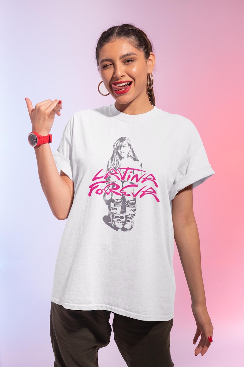 Puede incluir: Camiseta blanca con un gr&aacute;fico rosa y gris. El gr&aacute;fico presenta una figura sentada y el texto "Latina Forever". La camiseta la lleva una persona con cabello oscuro y un reloj rojo.