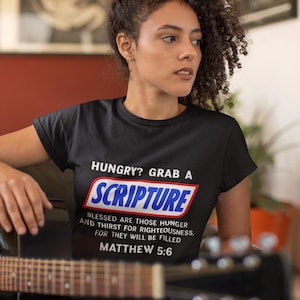 May include: Black t-shirt with the text "HUNGRY? GRAB A SCRIPTURE" in a candy bar-style design. The shirt also includes a Bible verse from Matthew 5:6. A black acoustic guitar is in the foreground.