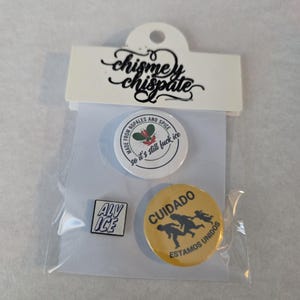 May include: A clear plastic package containing a set of four pins. The pins include a white pin with text, a yellow pin with figures and text, and a small square pin with text. The packaging has a white tag with black script.