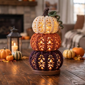 Puede incluir: Una luz decorativa apilada con tres calabazas de ganchillo en crema, naranja y morado oscuro, cada una con un intrincado patrón calado. Las calabazas están iluminadas desde el interior y se asientan sobre una base de madera con una enredadera.