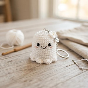 May include: A white crocheted ghost keychain featuring black bead eyes, a stitched smile, and pink blush accents. A small white flower is attached to the top, along with a silver metal ring and a circular charm engraved with the word Boo!.