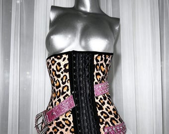 May include: A leopard print corset featuring a black central panel with hook-and-eye closures. The garment includes two pink glittery straps adorned with clear rhinestones, each secured by a silver-toned metal buckle on the front side.