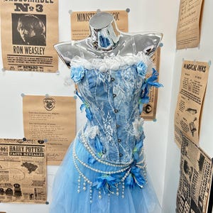 May include: A blue denim corset top featuring white lace overlays, a ruffled neckline, and blue floral accents. The corset is paired with a layered light blue tulle skirt adorned with draped pearl strands and blue flower embellishments.