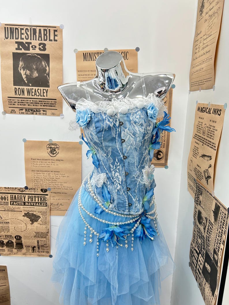 May include: A blue denim corset top featuring white lace overlays, a ruffled neckline, and blue floral accents. The corset is paired with a layered light blue tulle skirt adorned with draped pearl strands and blue flower embellishments.