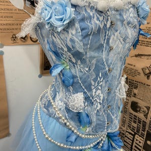 May include: A light blue corset top featuring white lace overlays, artificial light blue roses, and blue flower petals. The garment includes silver-toned front clasps and is adorned with multiple strands of white faux pearl beads.