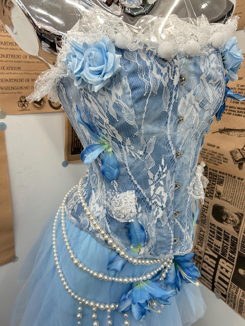 May include: A light blue corset top featuring white lace overlays, artificial light blue roses, and blue flower petals. The garment includes silver-toned front clasps and is adorned with multiple strands of white faux pearl beads.