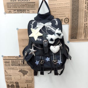 May include: A black backpack with a subtle plaid pattern, featuring a white lace bow, a plush star, a fuzzy black and white charm, a heart-shaped pendant, and silver-toned star charms hanging from chains. The bag has a flap closure with a buckle.