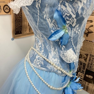 May include: A light blue corset top featuring white lace overlays, adorned with blue fabric flowers at the neckline and waist. The garment is embellished with draped strands of white faux pearls that wrap around the bodice and waistline.