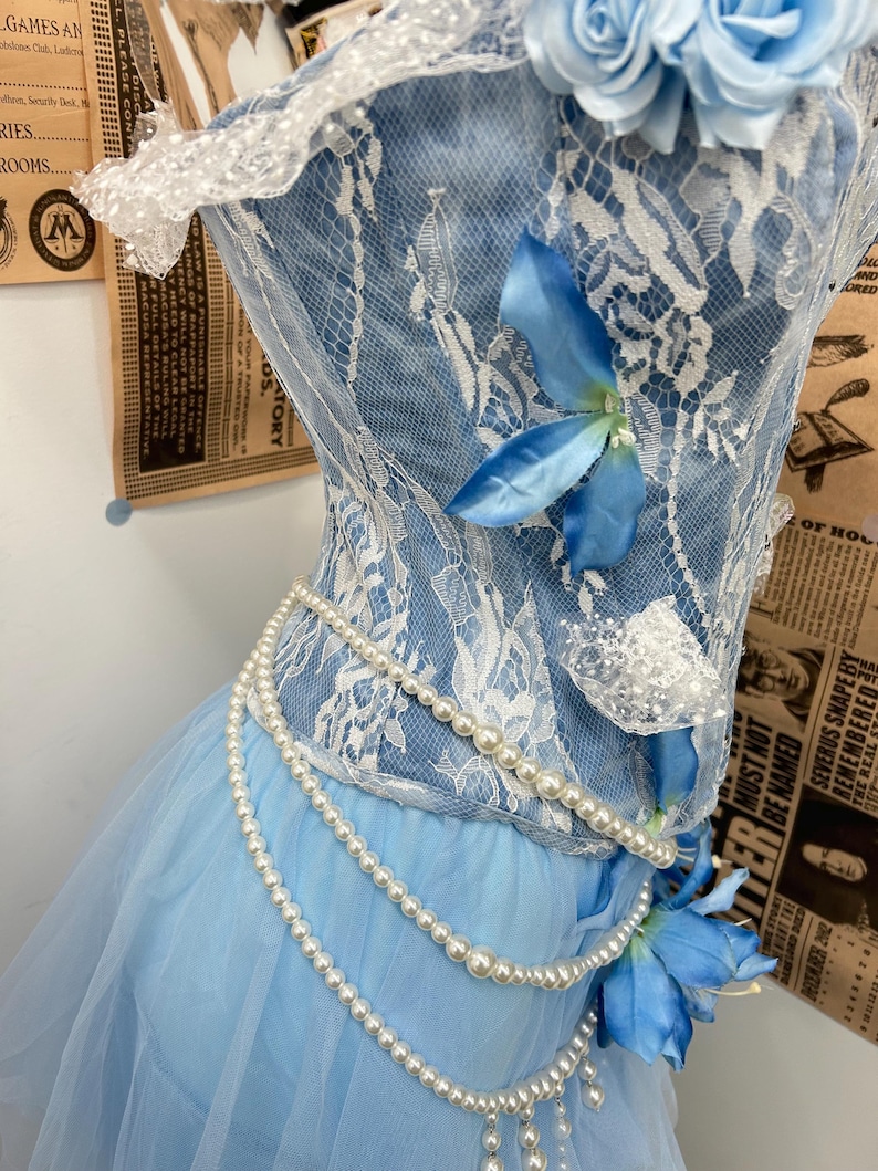 May include: A light blue corset top featuring white lace overlays, adorned with blue fabric flowers at the neckline and waist. The garment is embellished with draped strands of white faux pearls that wrap around the bodice and waistline.