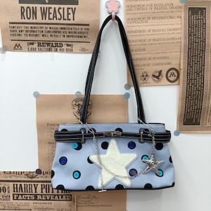 May include: A light blue shoulder bag featuring a large white star appliqué, black polka dots, and scattered blue buttons. The bag includes a black strap, a horizontal black belt-like accent with a silver clasp, and a silver star-shaped charm.