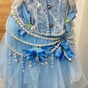 May include: A light blue corset top with white lace overlay paired with a layered tulle skirt. The ensemble is decorated with strings of white faux pearls, blue fabric flower petals, and two large blue artificial lily blossoms.