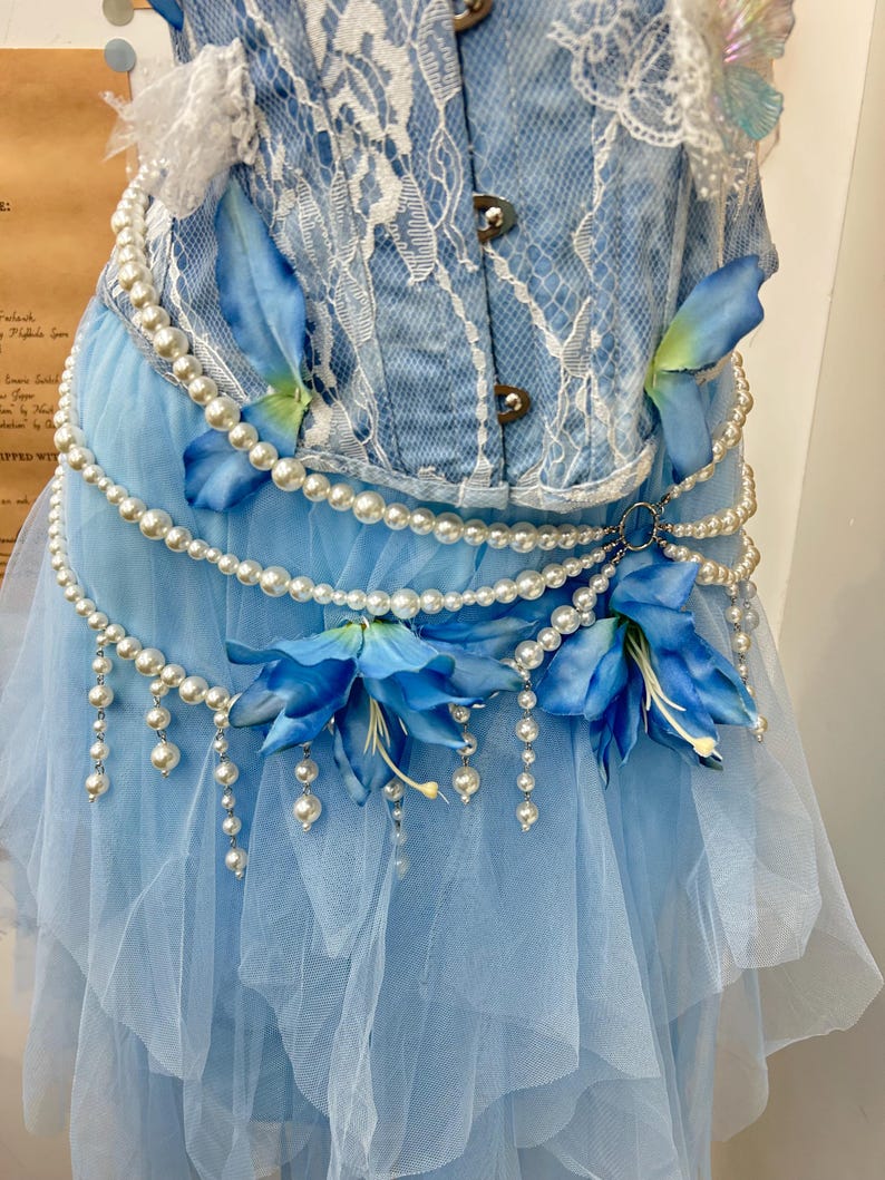 May include: A light blue corset top with white lace overlay paired with a layered tulle skirt. The ensemble is decorated with strings of white faux pearls, blue fabric flower petals, and two large blue artificial lily blossoms.