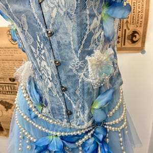 May include: A light blue denim corset featuring white lace overlays, silver metal front clasps, and iridescent butterfly accents. The garment is adorned with strings of white faux pearls and blue fabric flowers at the waistline.
