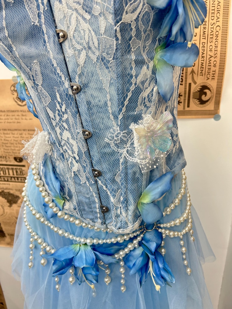 May include: A light blue denim corset featuring white lace overlays, silver metal front clasps, and iridescent butterfly accents. The garment is adorned with strings of white faux pearls and blue fabric flowers at the waistline.