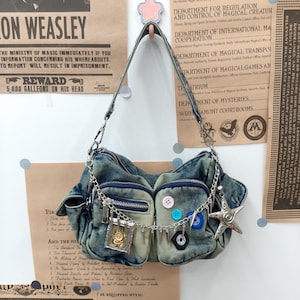 May include: A blue acid-wash denim shoulder bag featuring two front pockets with zippers, a silver chain strap, and decorative accents including colorful buttons, a small framed charm, and a large silver-toned star-shaped pendant.