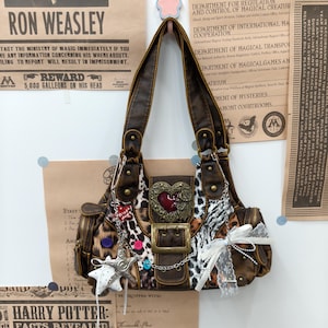 May include: A brown faux leather shoulder bag featuring a mix of leopard and zebra animal prints, a central heart-shaped emblem, a large buckle, and decorative charms including a silver star, white lace ribbons, and small colorful buttons.