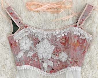 May include: A sleeveless corset top featuring a pink and red floral tapestry pattern, adorned with white floral lace appliqués, sequins, and beads. The garment includes a white ruffled hem, a scalloped lace neckline, and matching pale pink ribbon ties.