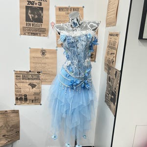 May include: A light blue corset top with white lace overlays and blue floral accents, paired with a layered tulle skirt featuring hanging pearl strands and blue flower embellishments, displayed on a chrome mannequin torso.