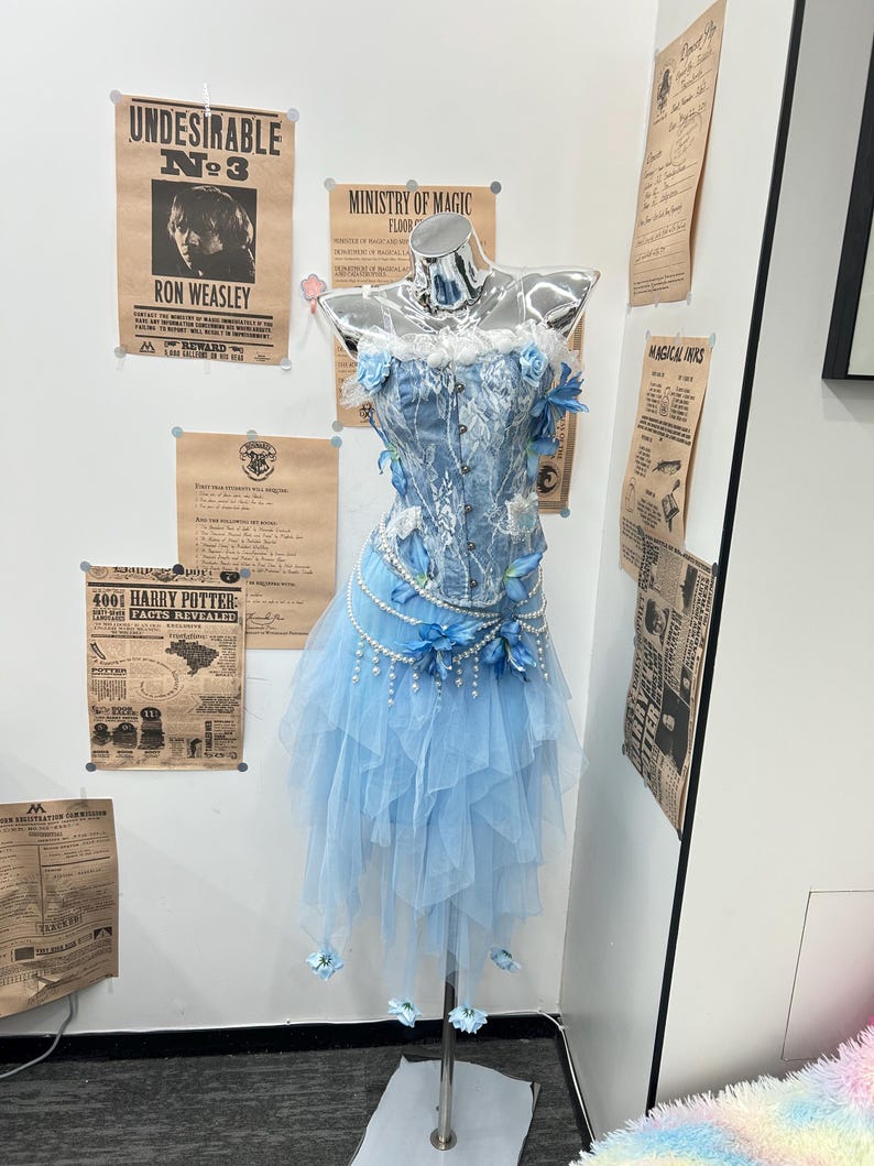 May include: A light blue corset top with white lace overlays and blue floral accents, paired with a layered tulle skirt featuring hanging pearl strands and blue flower embellishments, displayed on a chrome mannequin torso.