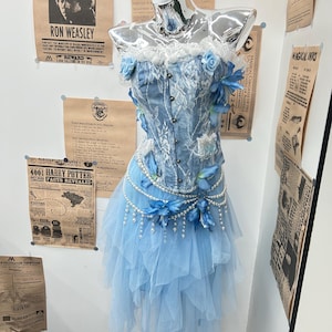 May include: A light blue corset top with white lace overlays and a front button closure, paired with a layered tulle skirt. The ensemble is decorated with blue fabric flowers and draped strings of white faux pearls at the waistline.