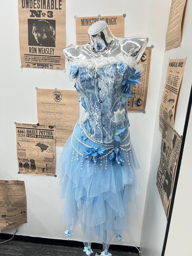 May include: A light blue corset top with white lace overlays and a front button closure, paired with a layered tulle skirt. The ensemble is decorated with blue fabric flowers and draped strings of white faux pearls at the waistline.