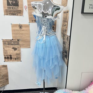 May include: A light blue corset dress featuring a lace-up back with white ribbons, a layered tulle skirt, and pearl-like beaded accents. The bodice has white lace trim along the top edge, and the skirt is composed of multiple asymmetrical tulle layers.