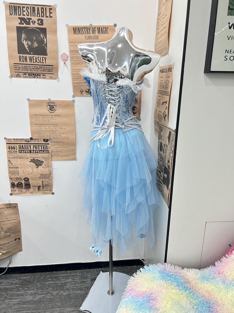 May include: A light blue corset dress featuring a lace-up back with white ribbons, a layered tulle skirt, and pearl-like beaded accents. The bodice has white lace trim along the top edge, and the skirt is composed of multiple asymmetrical tulle layers.